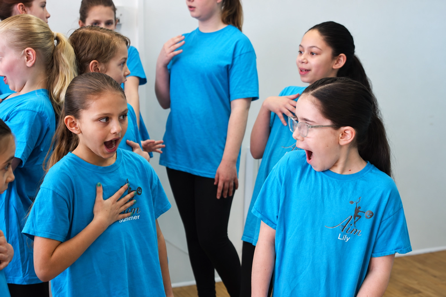 Students in turquoise AIM T-shirts practising singing and expressive gestures during a musical theatre class at Artists In Motion in Aylesham